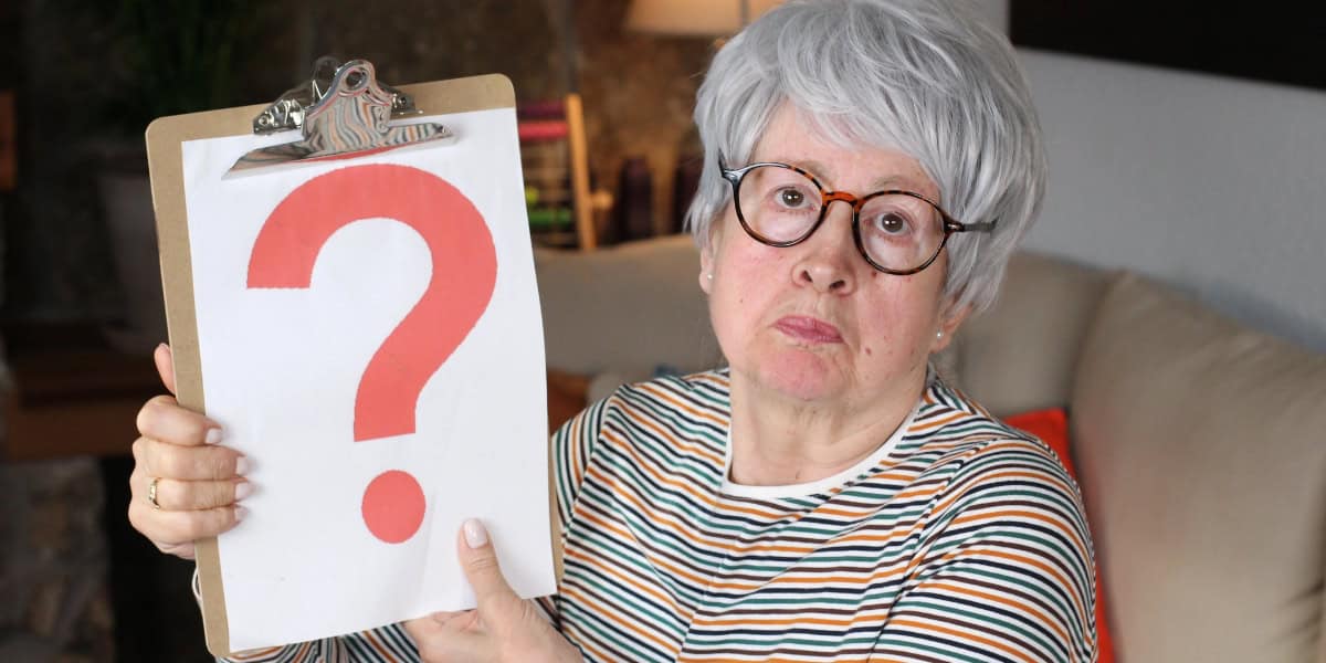 An older woman with gray hair and glasses holds up a clipboard displaying a large red question mark, sitting indoors on a couch.